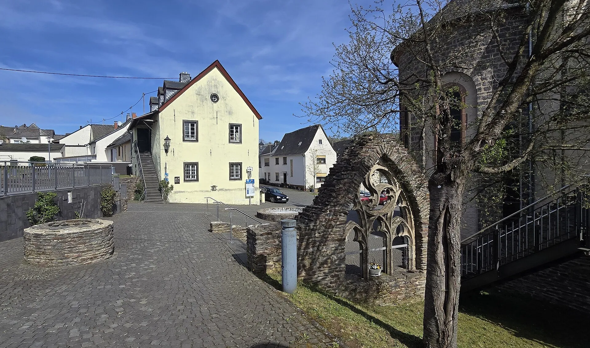 Gepflasterter Platz an der Burgstraße in Kaisersesch mit gelbem Giebelhaus, Brunnenring und gotischem Maßwerkbogen