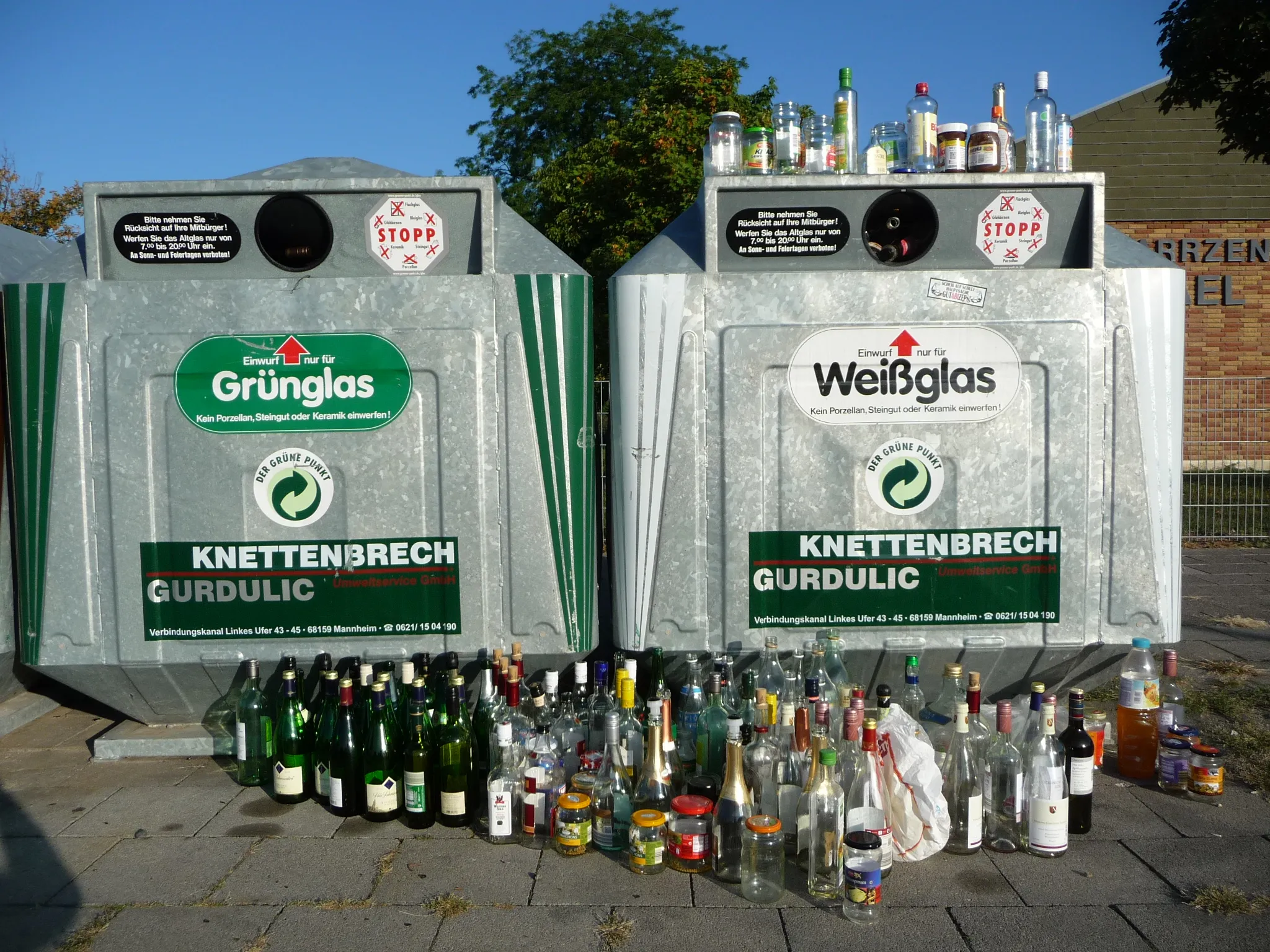 Zwei verzinkte Altglascontainer für Grün- und Weißglas, davor und darauf dicht gedrängt abgestellte leere Flaschen und Gläser